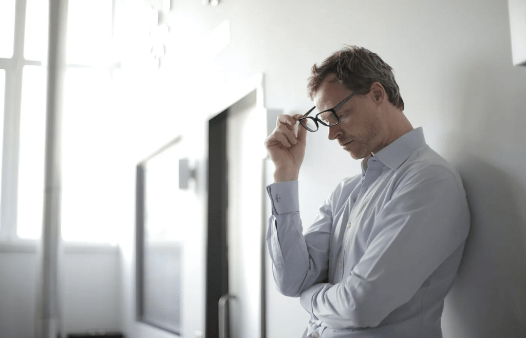 a man in a dress shirt stands against a wall, removing his glasses with his eyes closed, a look of weariness or stress on his face
